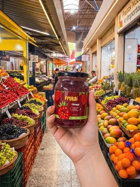 Mão segurando um pote de Frutaí no meio de uma feira de frutas